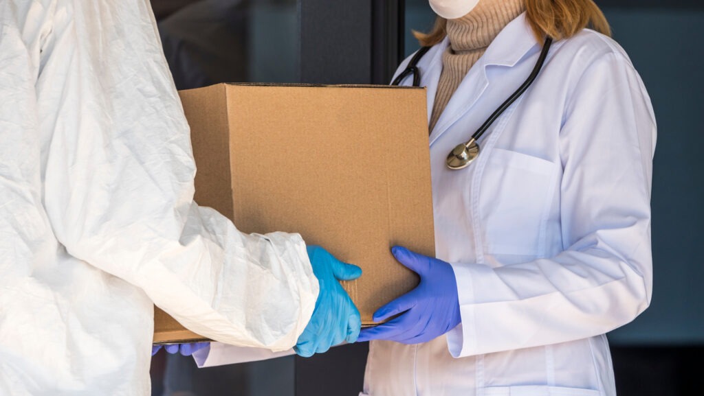 A medical professional in a white coat and mask accepts a cardboard box from a person in protective clothing, emphasizing healthcare delivery.