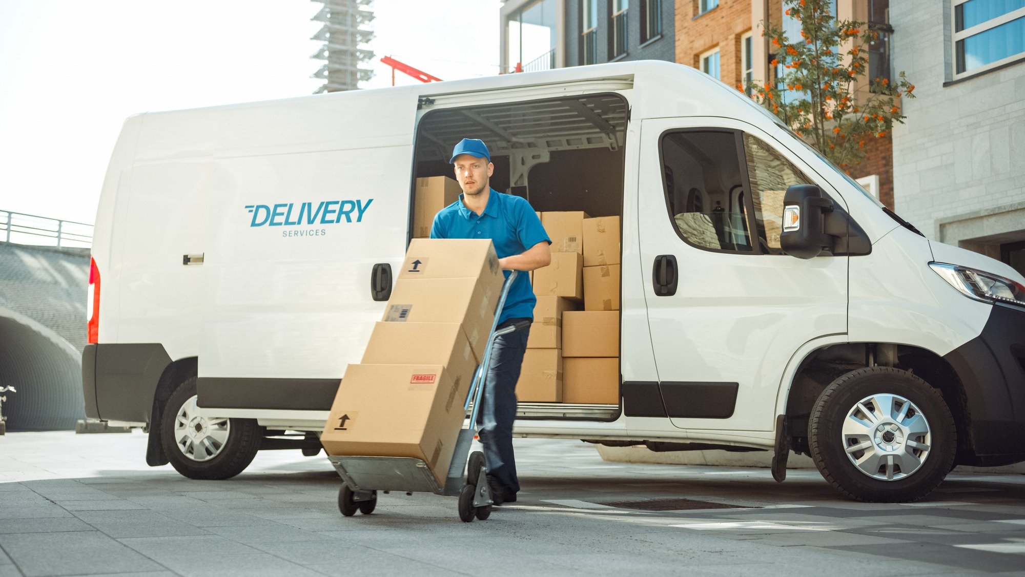 Delivery worker transporting boxes from a van labeled "DELIVERY SERVICES" in an urban area, showcasing logistics and shipment activities.